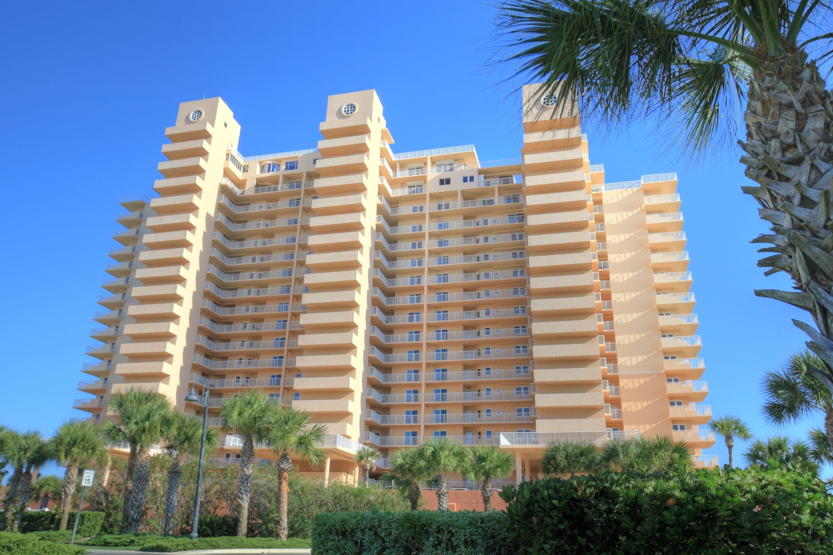 Un hôtel à plusieurs étages doté de balcons, de palmiers et surplombant un ciel d’un bleu limpide.