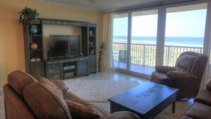 A living room with a television, brown leather sofas, a coffee table, and a view of the beach through large windows.