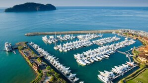 Una vista aerea di una marina con numerose imbarcazioni ormeggiate, circondata da una cittadina costiera con edifici e un molo che si protende nell'acqua.