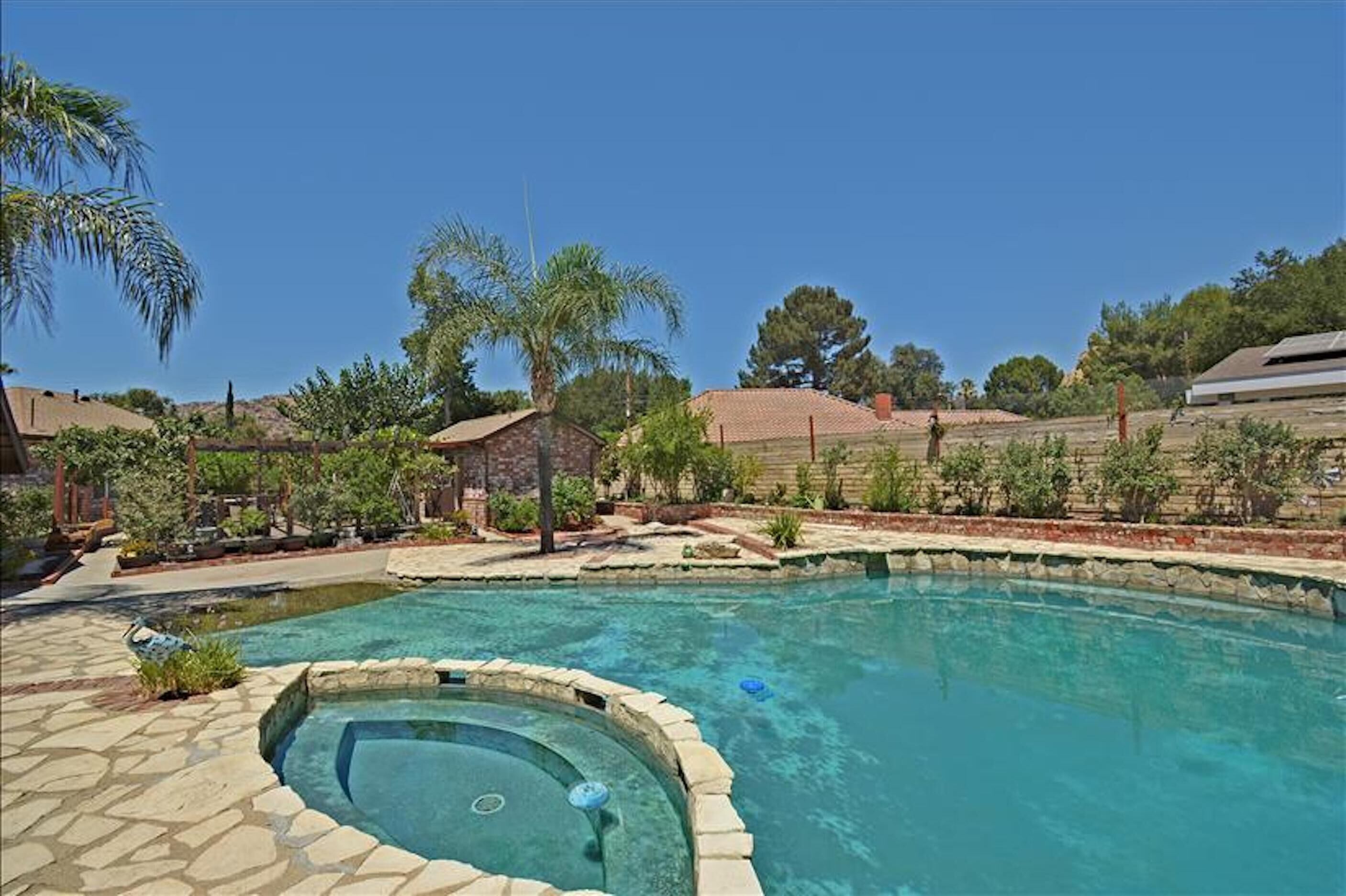 A swimming pool with a hot tub, surrounded by a landscaped garden with palm trees and a stone wall.