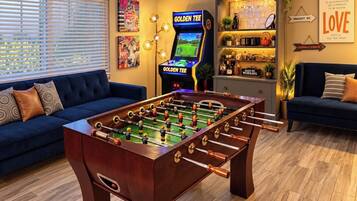 A room with a foosball table, a foosball table with a screen, and a bar area with shelves.