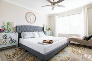 A bedroom with a grey upholstered bed, a ceiling fan, a window with blinds, a nightstand with a lamp, and a decorative wall piece. - Luxury 3-Bedroom Home with Pool Near Stadium-Perfect for Game Weekends (Glendale)