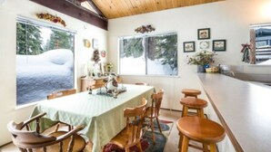 A dining area with a table set for four, wooden chairs, a bar area, and large windows with a view of trees and snow outside.