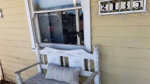 A rustic wooden bench with a cushion, positioned outside a house with a window and a number plaque on the wall.