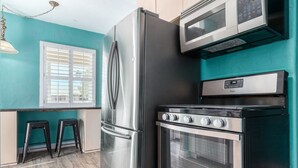 A kitchen with a stainless steel refrigerator, a microwave, and an oven. There are two black stools and a window with white shutters.