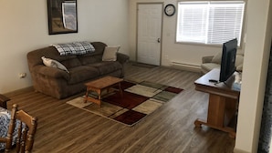 A living room with a brown sofa, a wooden coffee table, a rug, a television on a stand, and a door with a clock. - Tidy Country Cottage 5 minutes outside of Cody (Cody)