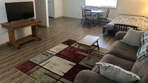 A living room with a TV, a wooden coffee table, a patterned rug, a sofa, and a dining area with chairs and a table. - Tidy Country Cottage 5 minutes outside of Cody (Cody)