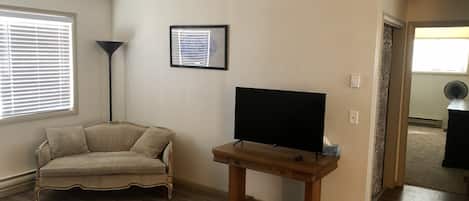 En stue med trægulv, en beige sofa, et fjernsyn på et træskab og en gulvlampe.