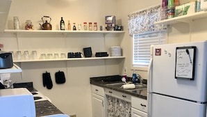 A compact kitchen with white cabinets, a black countertop, and a refrigerator. There are shelves above the counter holding various kitchen items, and a window with blinds in the background. - Tidy Country Cottage 5 minutes outside of Cody (Cody)