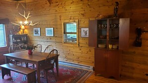 Un interior de madera con una mesa de comedor, sillas, una araña, cuadros enmarcados, un aparador con vajilla y una ventana con persianas.