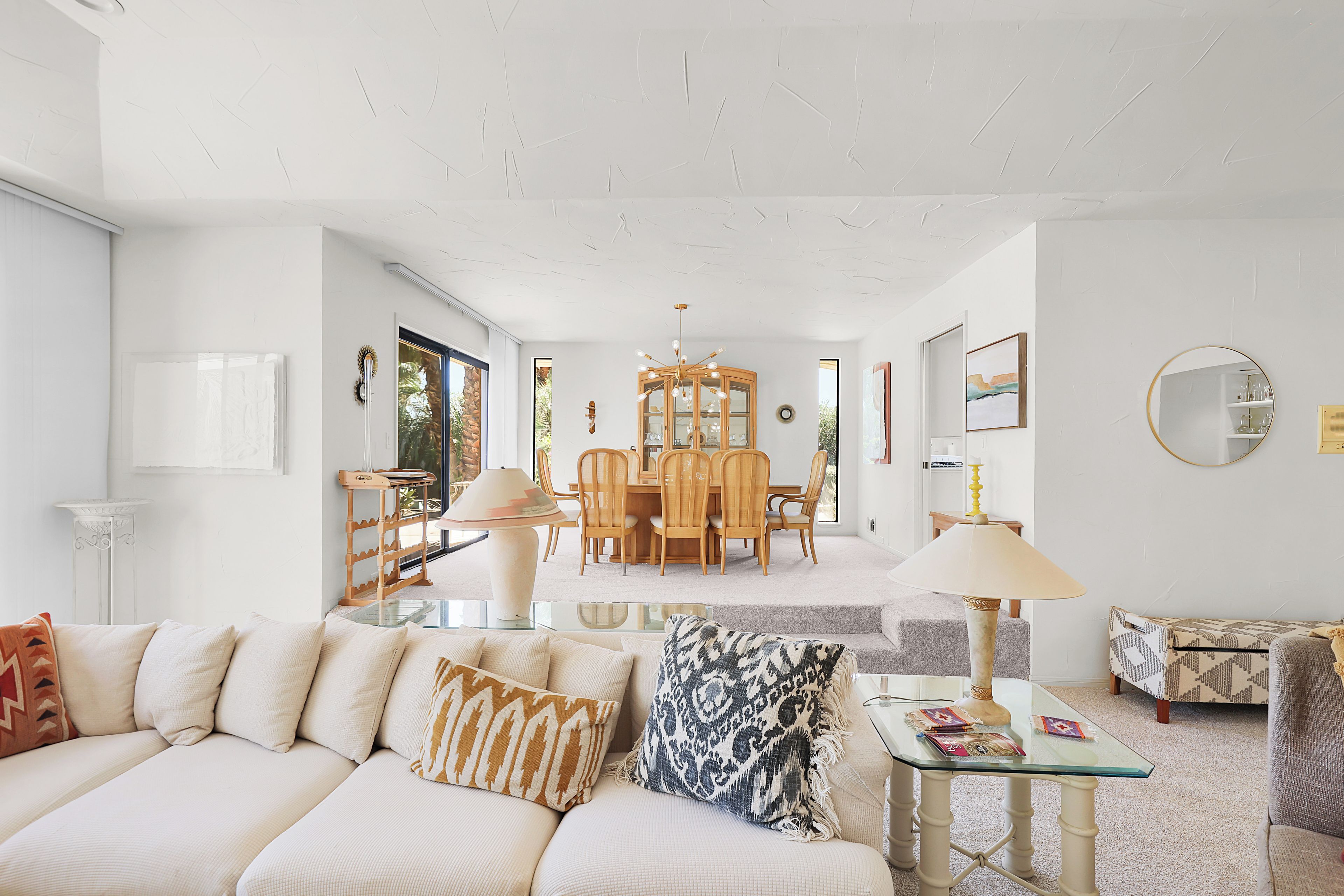 A spacious living room with a sofa, a glass coffee table, and a dining area with wooden chairs and a chandelier.