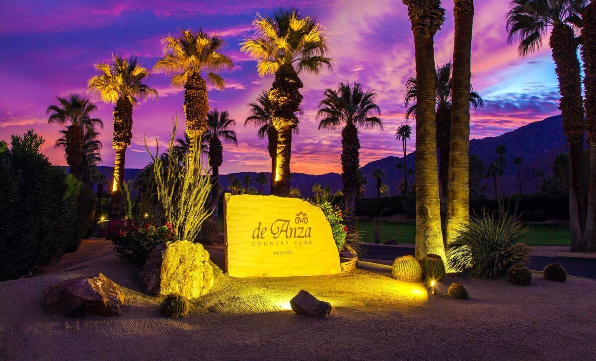 A private country club sign with palm trees and desert plants around it.