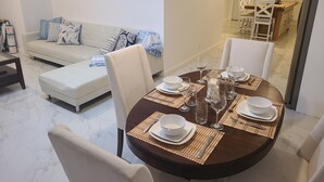 A dining area with a round table set for four, a sofa with patterned cushions, and two framed paintings on the wall.