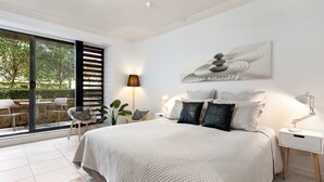 A modern bedroom with a large bed, a side table, a lamp, and a view of the outdoors through a sliding door. - Harbourside Studio Apartment (Kiama)