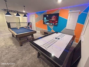 A room with a pool table, a foosball table, and a TV mounted on a colorful wall.