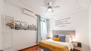 A hotel room with a bed, a nightstand, a lamp, a ceiling fan, a window with curtains, and a wall with a cityscape mural. - vip  LETRAS, by MONARO***** (Madrid)