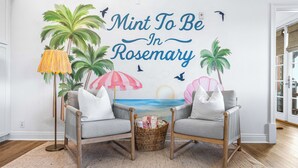 Una habitación con dos sillones grises, una cesta de mimbre y un mural en la pared con palmeras y una playa.