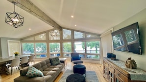 A spacious living area with a large sofa, ottomans, and a dining table. There are large windows offering views of the outdoors. - Gorgeous Waterfront home with sweeping views of a beautiful bay. (Bath)