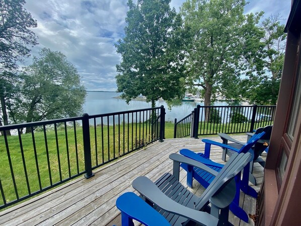 A wooden deck with a railing overlooking a lake, featuring blue Adirondack chairs and a grassy area. - Gorgeous Waterfront home with sweeping views of a beautiful bay. (Bath)