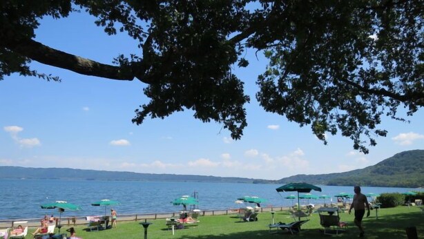 Eine Terrassenausstattung am See mit Liegestühlen und Sonnenschirmen, eine Rasenfläche und Blick auf See und Hügel.