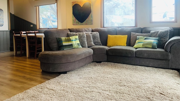 A living room with a sectional sofa, a dining table with chairs, a large window, and a heart-shaped wall decoration.