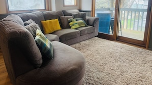 A living room with a sectional sofa, a large window, and a sliding glass door leading to a deck.