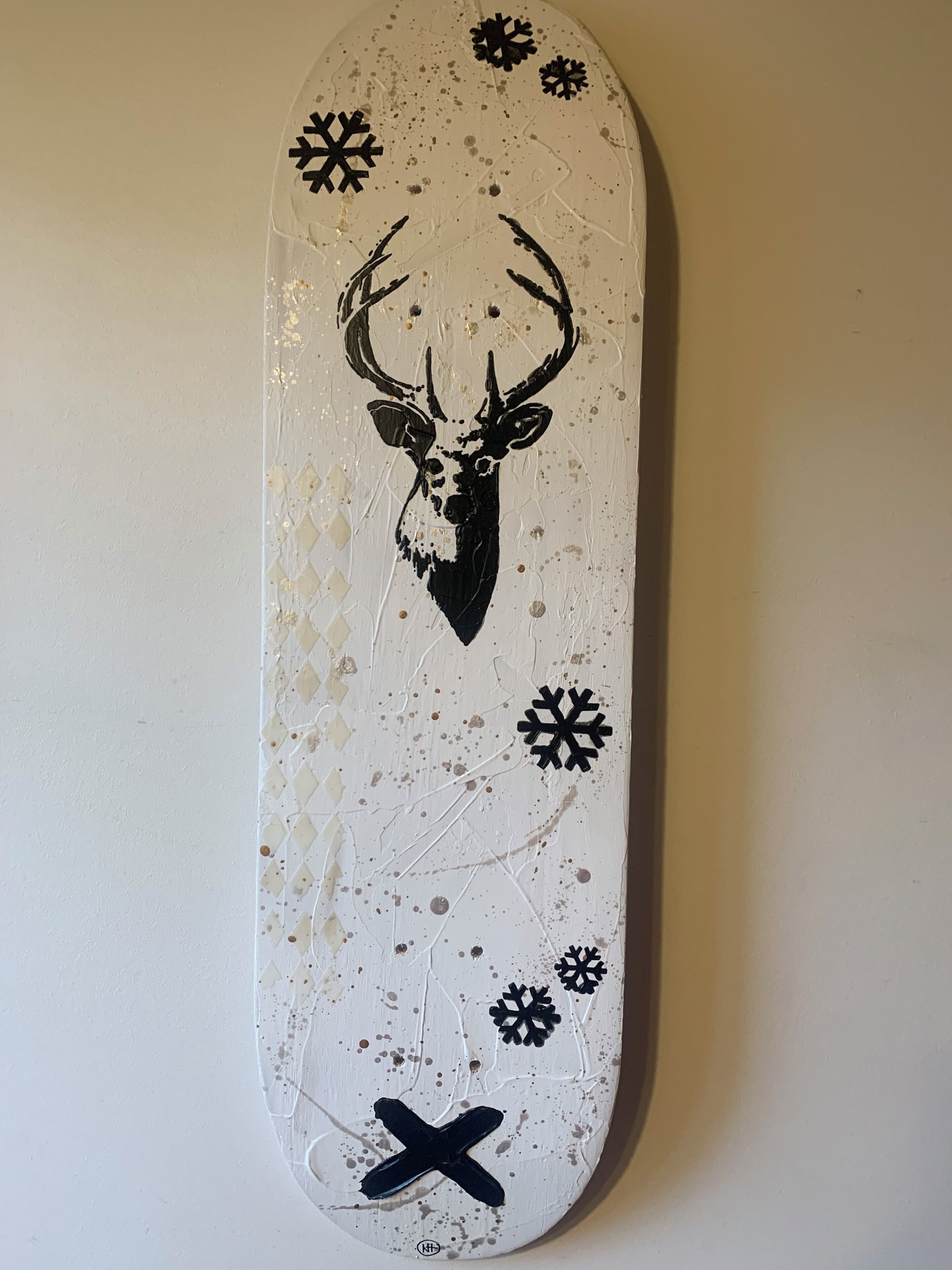Un monopatín con diseño de cabeza de ciervo, adornado con copos de nieve y una 'X' negra en la parte inferior.