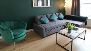 A modern living room with a green armchair, grey sofa with teal cushions, and a glass coffee table with a plant. - Sea Nest (Torquay)