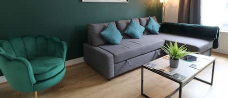 A modern living room with a green armchair, grey sofa with teal cushions, and a glass coffee table with a plant.