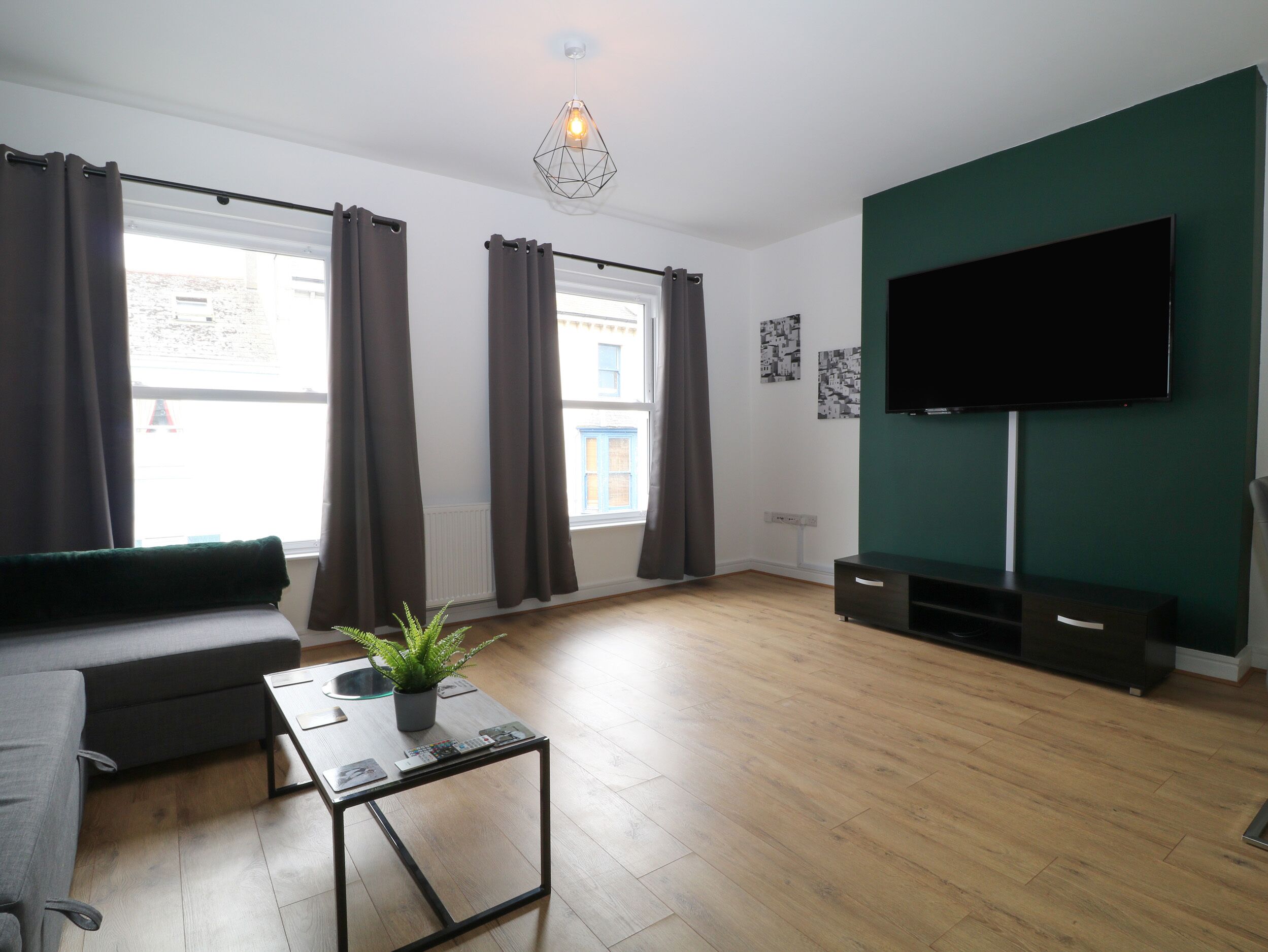 Una sala moderna con un sofá, una mesa de centro con una planta, un televisor de pantalla plana y un ventanal con cortinas.