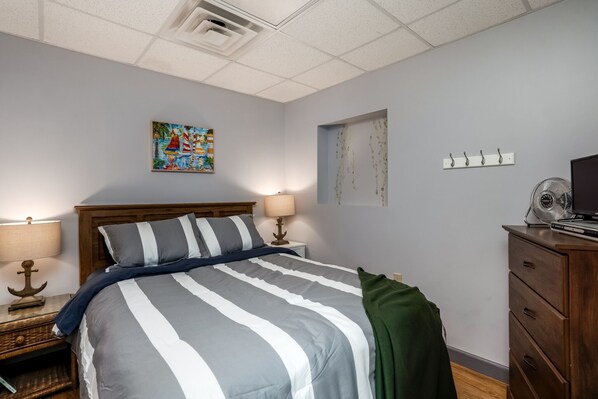Une chambre à coucher bien rangée, avec un lit, deux lampes de chevet, une petite table surmontée d’une lampe en forme d’ancre, un ventilateur fixé au mur et une commode.