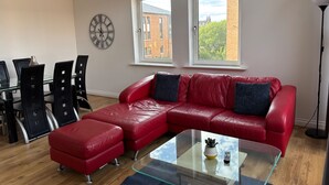 A modern living room with a red leather sofa, a glass coffee table, a dining table with chairs, and a large wall clock. - The OVO Hydro Penthouse With Free Parking (Glasgow)