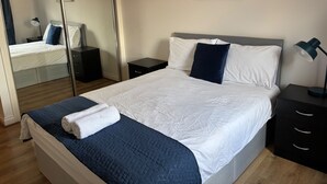 A neatly made bed with white and navy bedding, a dark blue quilt, and a white towel. There is a painting of a forest on the wall, and a mirror reflecting the room. - The OVO Hydro Penthouse With Free Parking (Glasgow)
