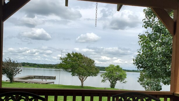 Un espace extérieur aménagé, abrité et doté de tables et de chaises, offrant une vue sur un lac bordé d’arbres et sur un quai.
