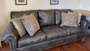 Un sofá de cuero marrón oscuro con costuras decorativas y dos almohadones estampados, situado frente a una pared blanca con cuadros enmarcados y un reloj de pared.