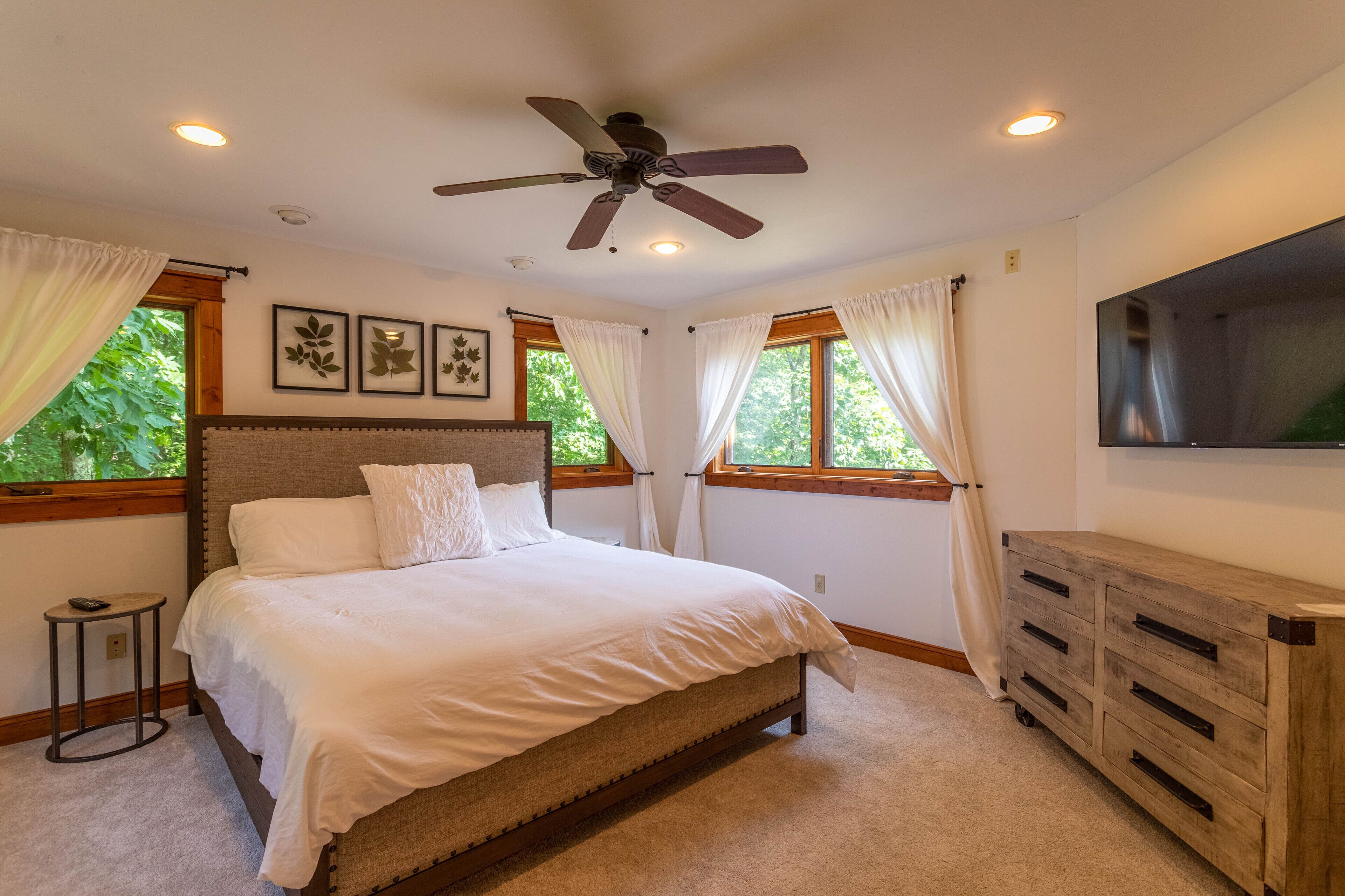 A bedroom with a bed, ceiling fan, large windows, and a TV.
