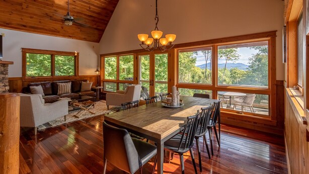 A spacious dining area with a wooden table, chairs, and a chandelier. Large windows offer views of trees and mountains. - Shady Pines - modern mountain home near winery, game table, fire pit (Banner Elk)