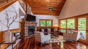 A spacious living room with a stone fireplace, leather sofas, a flat-screen TV, and large windows offering a view of the outdoors. - Shady Pines - modern mountain home near winery, game table, fire pit (Banner Elk)