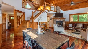 A spacious living area with a large wooden dining table, comfortable seating, a stone fireplace, and a staircase leading to an upper level. - Shady Pines - modern mountain home near winery, game table, fire pit (Banner Elk)