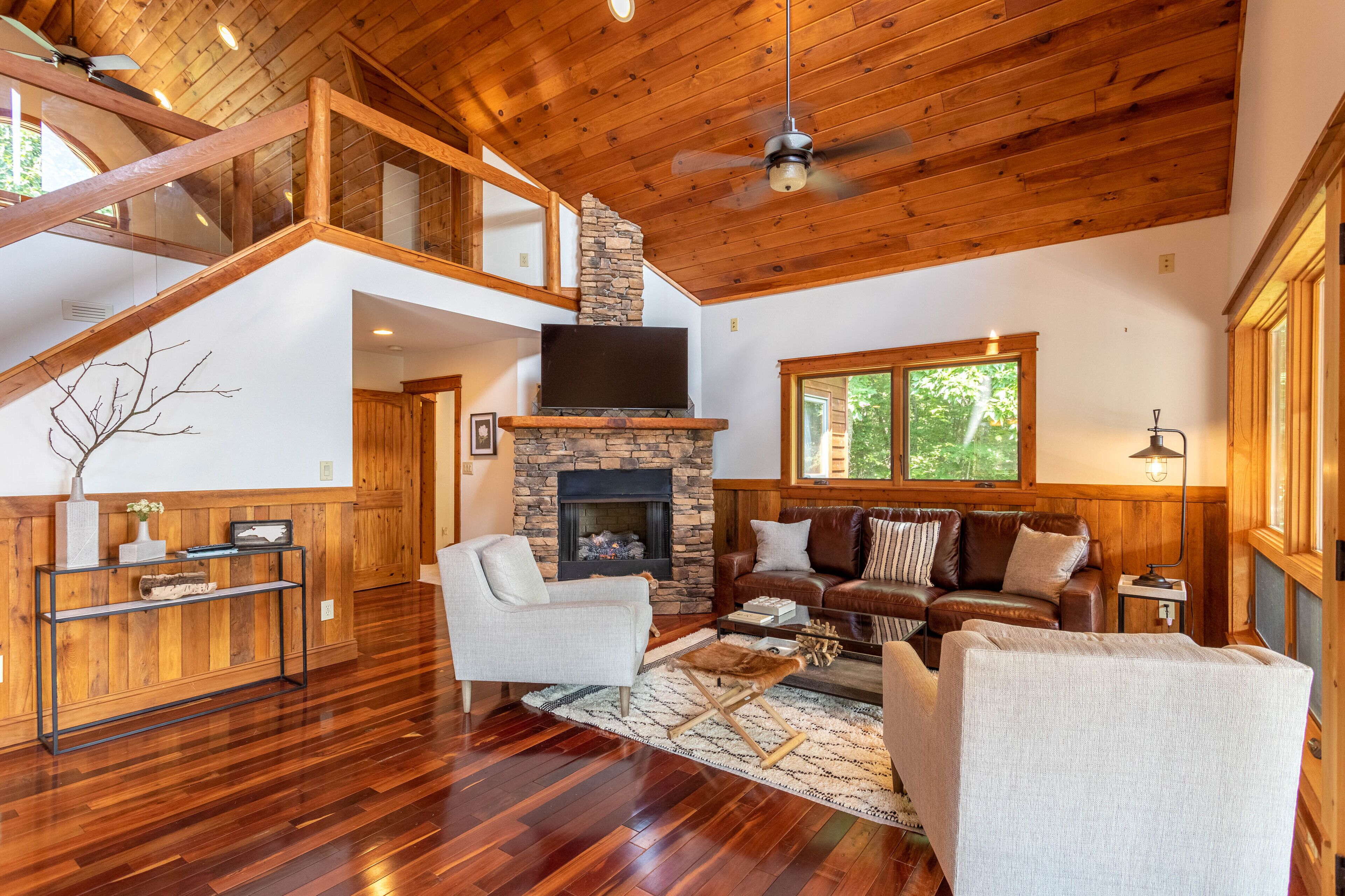 A spacious living area with a stone fireplace, wooden flooring, and a high ceiling with exposed beams.