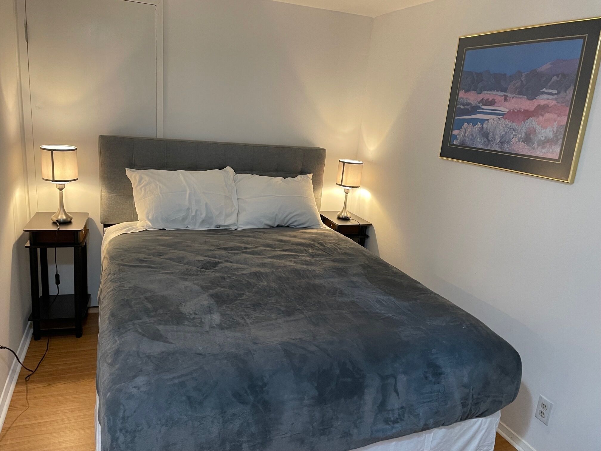 Ein ordentlich bezogenes Bett mit einer grauen Daunendecke, zwei Nachttischlampen und einem gerahmten Landschaftsbild an der Wand.