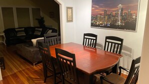 A dining area with a wooden table and chairs, a framed cityscape picture on the wall, and a living room in the background. - Olympic Hills six bed haven with hot tub and grill (Seattle)