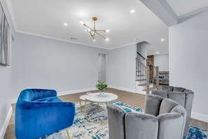 A modern living room with a blue armchair, grey sofas, a round coffee table, and a chandelier. - Modern Home w. Large Dining Area | Playset & WiFi (Suffern)