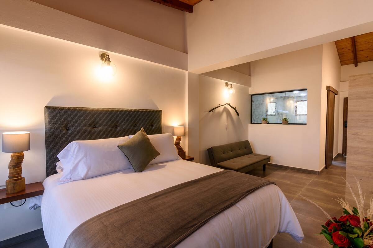 A hotel room with a bed, bedside lamps, a bench, a mirror, and a window.