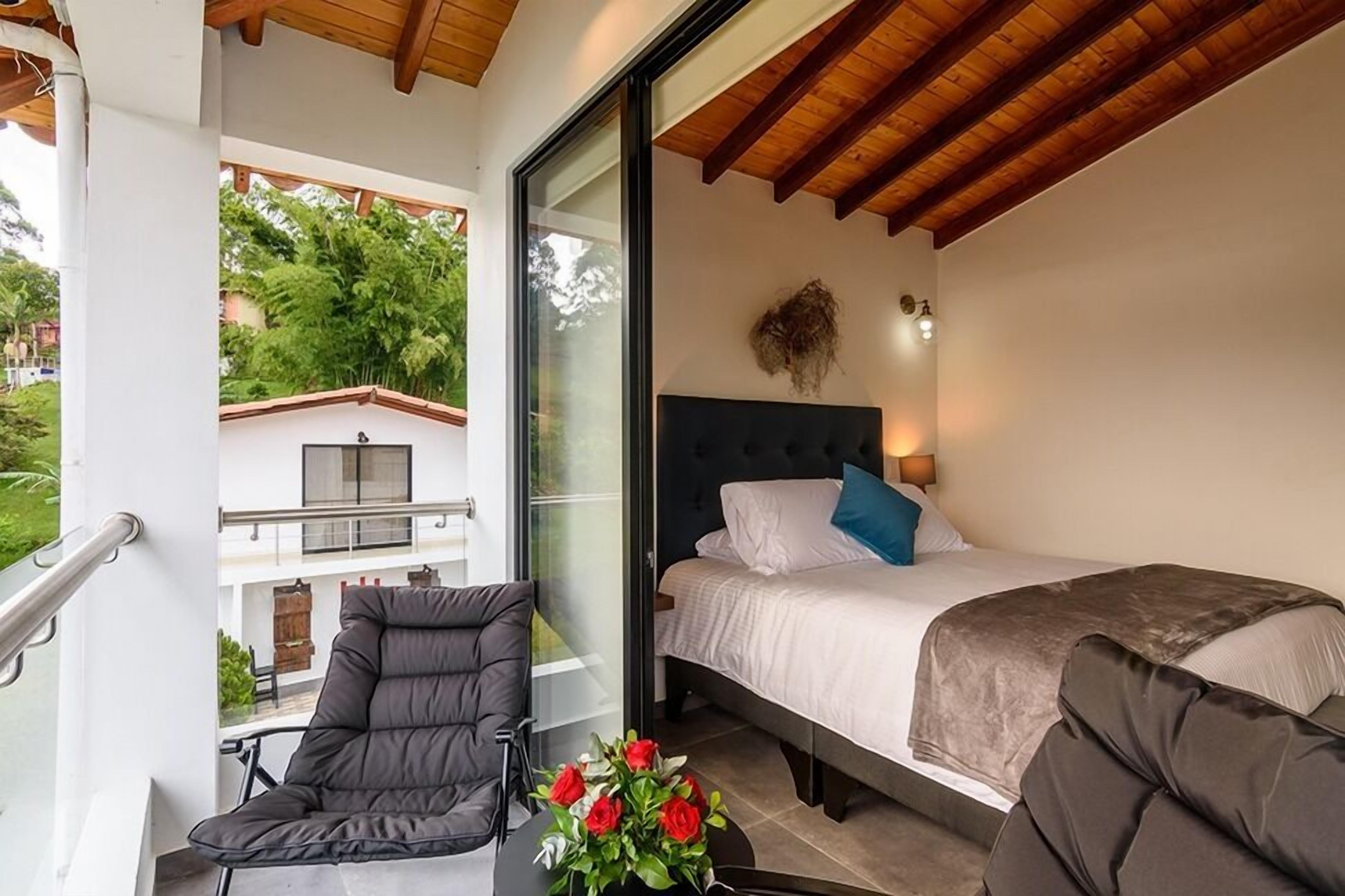 A bedroom with a bed, a chair, and a balcony with a view of greenery.