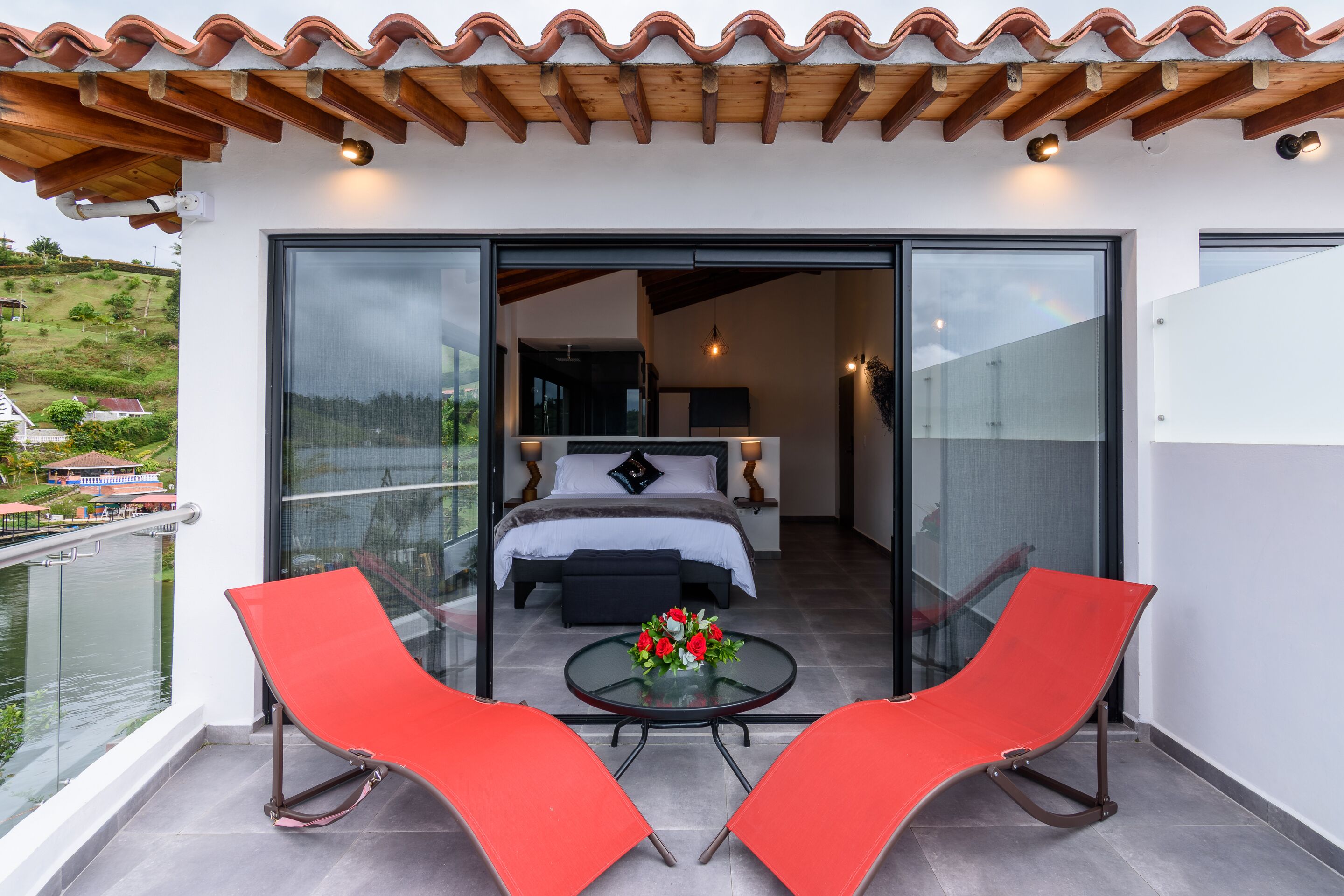 Un balcón con una cama, una mesa adornada con flores y dos sillas rojas.