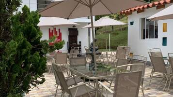 Un patio al aire libre con una mesa redonda y sillas, rodeado de sombrillas, y una casa con techo de tejas al fondo.