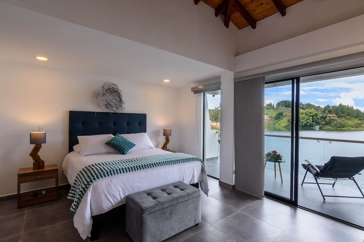 A modern bedroom with a large bed, a bench, and a view of a lake through a sliding glass door.