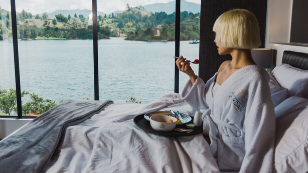 A person in a robe sitting on a bed with a tray of breakfast items, overlooking a lake with hills in the background. - Jordan Hotel Boutique (El Peñol)