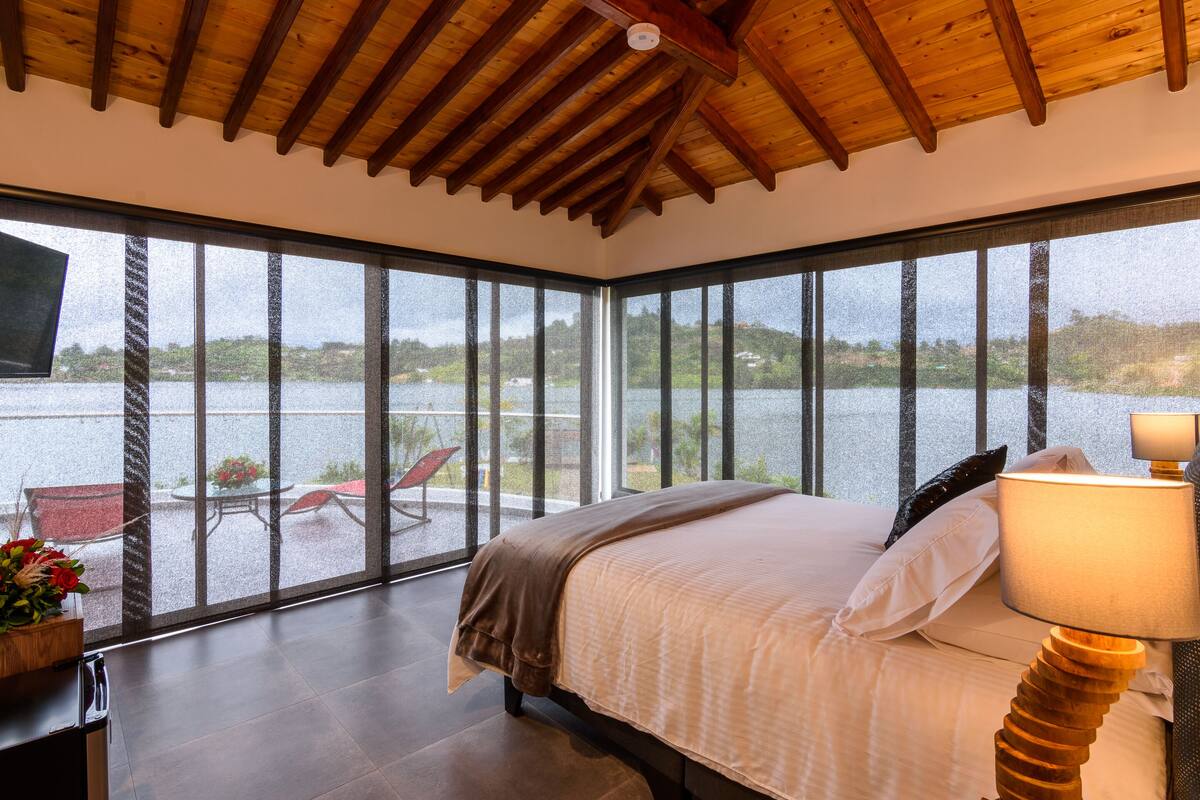 A bedroom with a large window overlooking a body of water, a bed with a wooden headboard, and a table lamp.